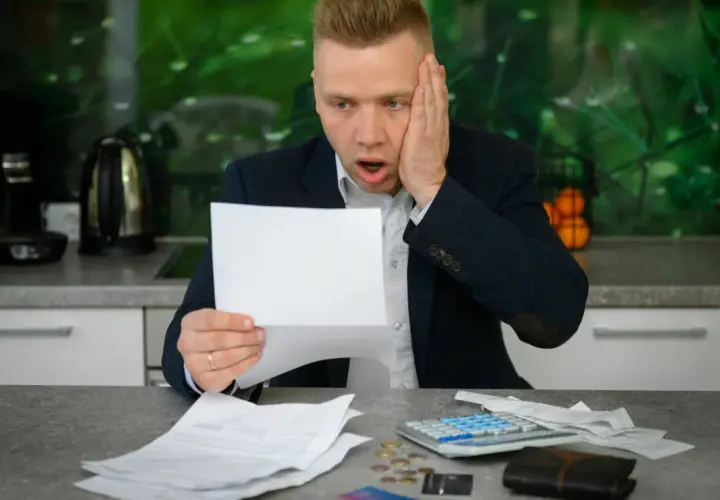 Zaskoczony mężczyzna trzymający rachunek, obok kalkulator i monety.