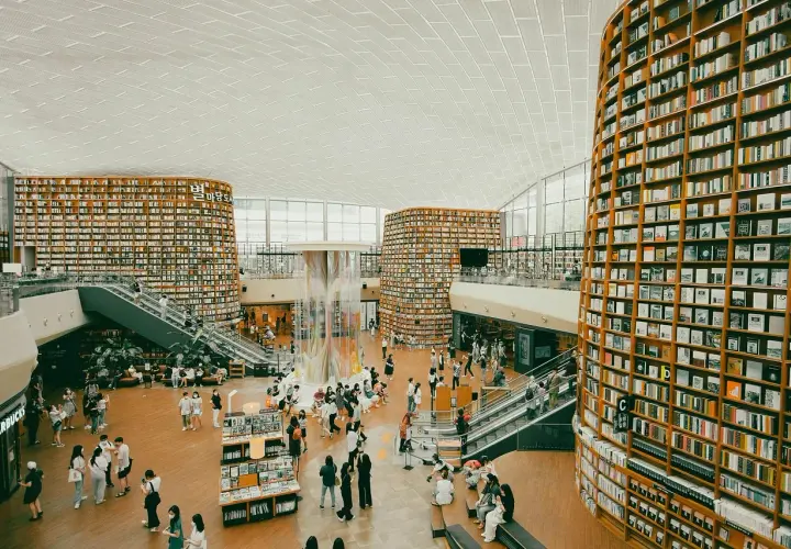 Wielopoziomowa biblioteka z półkami sięgającymi sufitu, gdzie każdy **bibliotekarz** marzyłby o pracy. Ludzie spacerują, czytają i odpoczywają wśród książek.
