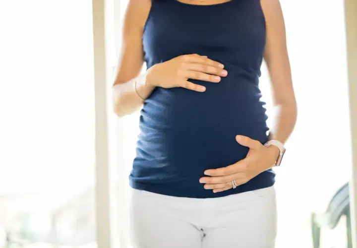 Ciężarna kobieta w granatowej bluzce i białych spodniach obejmuje dłońmi brzuch. Wody płodowe otulają rozwijające się życie.