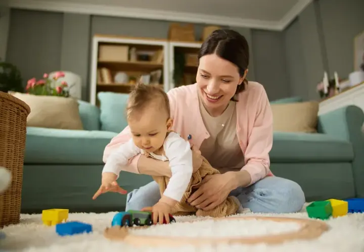 Uśmiechnięta mama bawi się z dzieckiem klockami i pociągiem na dywanie w salonie.