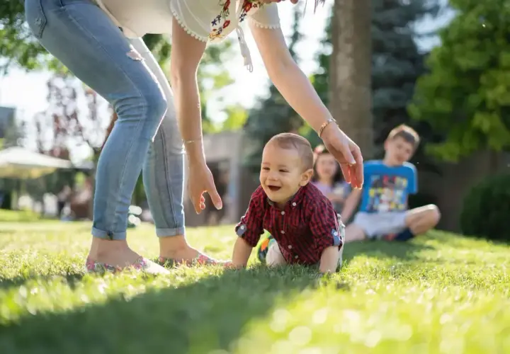 Uśmiechnięte dziecko raczkuje na trawie, a opiekunka wyciąga do niego ręce.