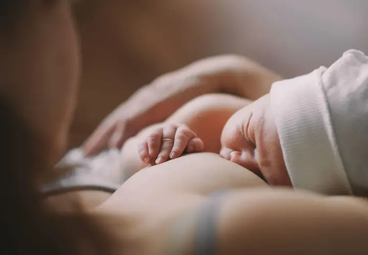 Delikatne dłonie noworodka obejmujące pierś podczas karmienia. Ulewanie u noworodka to częsty widok, ale ten moment to czysta bliskość.