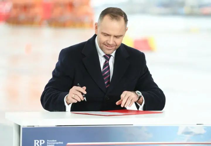 Mężczyzna w garniturze i płaszczu podpisuje dokumenty przy stole.