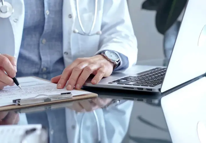 Lekarz w białym kitlu pisze na dokumentach, obok laptop.