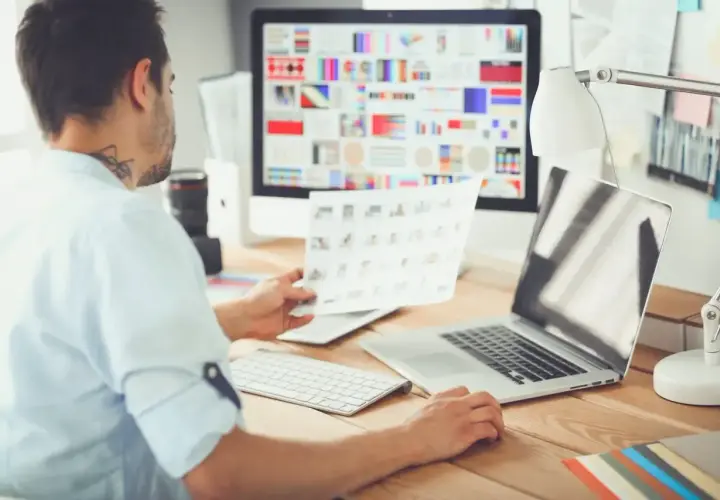 Mężczyzna przy biurku przegląda wydruki, przed nim komputer z kolorowymi grafikami i laptop.