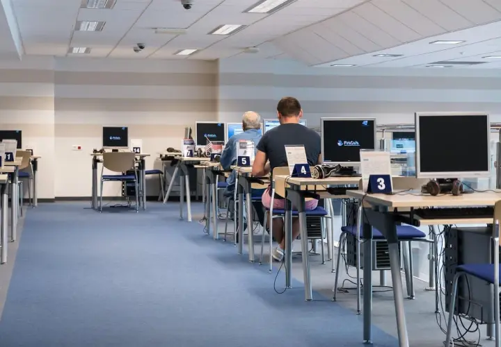 Rząd stanowisk komputerowych z osobami pracującymi, niebieska wykładzina.