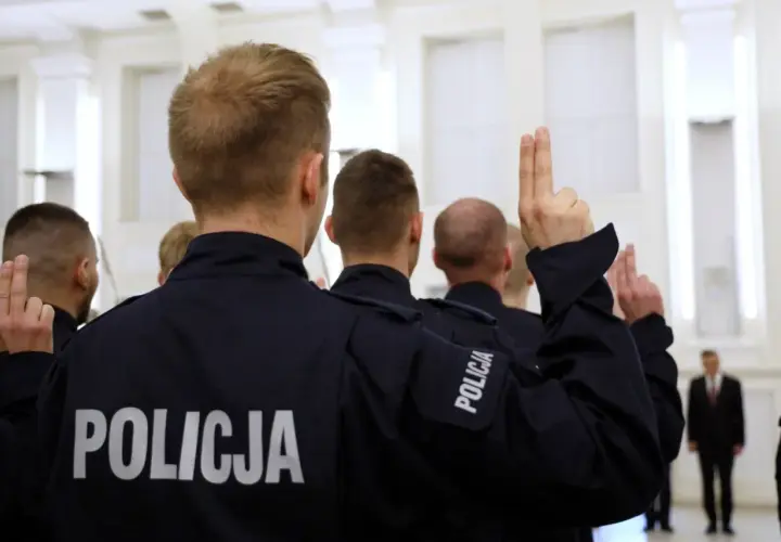 Policjanci w mundurach z napisem "POLICJA" podnoszą ręce z dwoma palcami uniesionymi do góry.