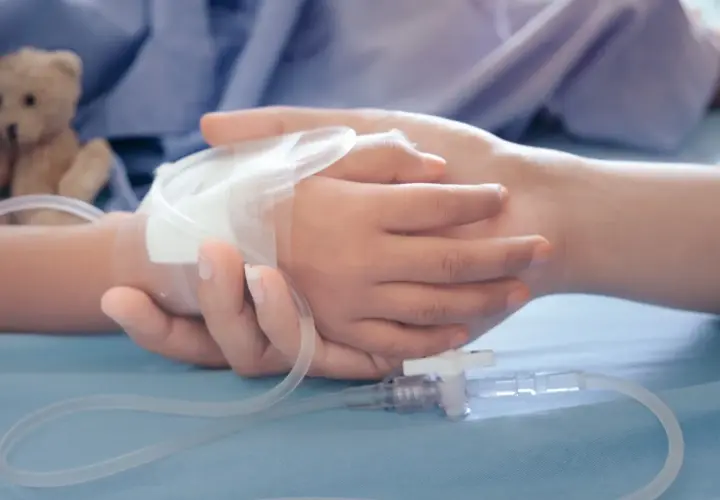 Dłoń dorosłego obejmuje dłoń dziecka z wenflonem. W tle pluszowy miś. Obraz symbolizuje wsparcie i troskę, jakiej wymaga zasilek pielegnacyjny.