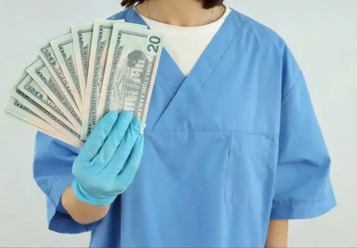 Higienistka stomatologiczna w niebieskim fartuchu trzyma wachlarz banknotów 20-dolarowych.