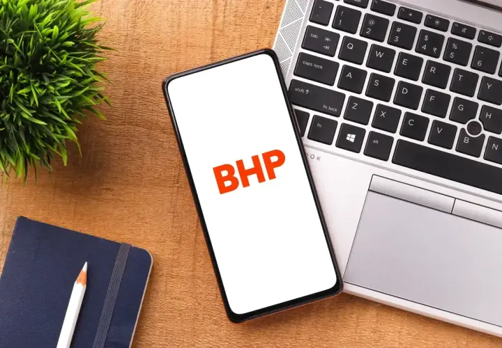 Telefon z napisem BHP na ekranie, obok laptopa, notatnika i rośliny.