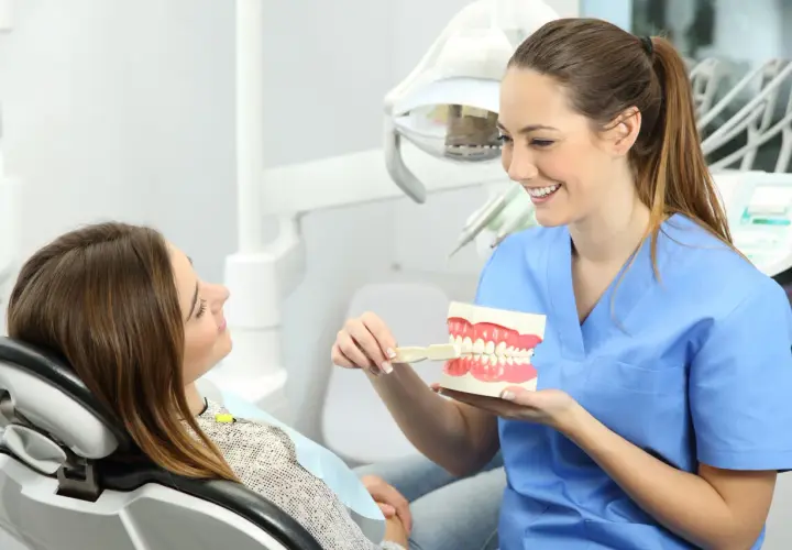Uśmiechnięta higienistka stomatologiczna pokazuje pacjentce model szczęki i szczoteczkę.