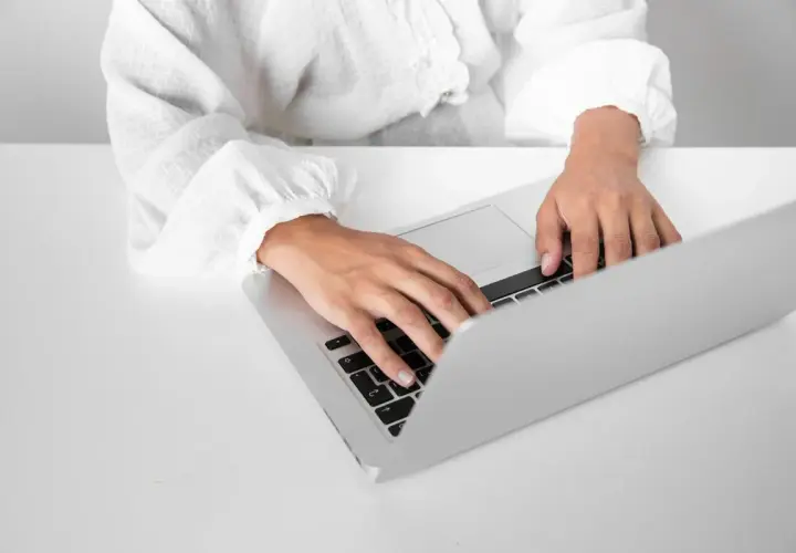 Dłonie piszące na klawiaturze laptopa na białym biurku.