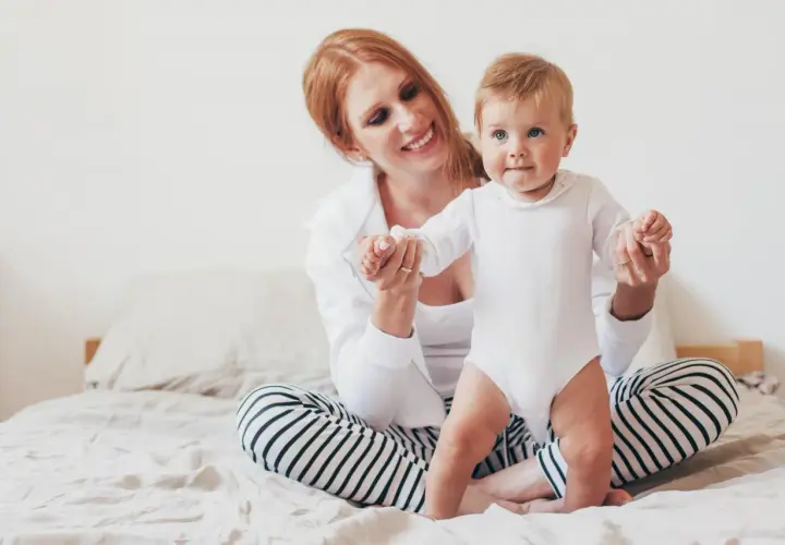 Uśmiechnięta mama z rudymi włosami pomaga 8-miesięcznemu dziecku w białym body wstać na łóżku.