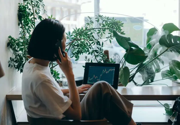 Kobieta rozmawia przez telefon przy laptopie z wykresem giełdowym, otoczona roślinami.