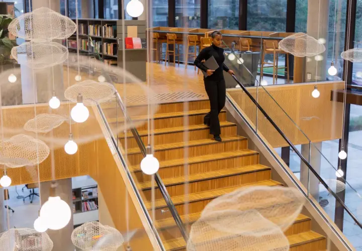 Kobieta z laptopem schodzi po drewnianych schodach w nowoczesnej bibliotece z ozdobnymi chmurkami i lampami.