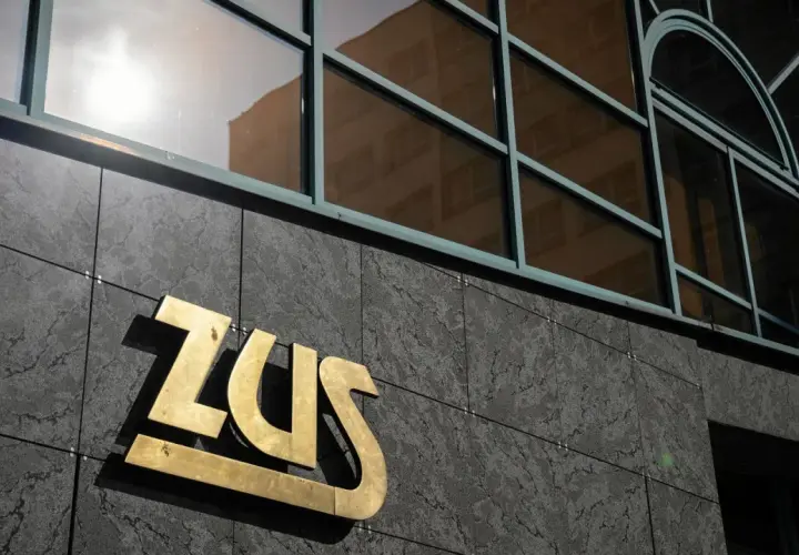 Złote logo ZUS na ciemnej, kamiennej elewacji budynku, nad nim okna z odbiciem.