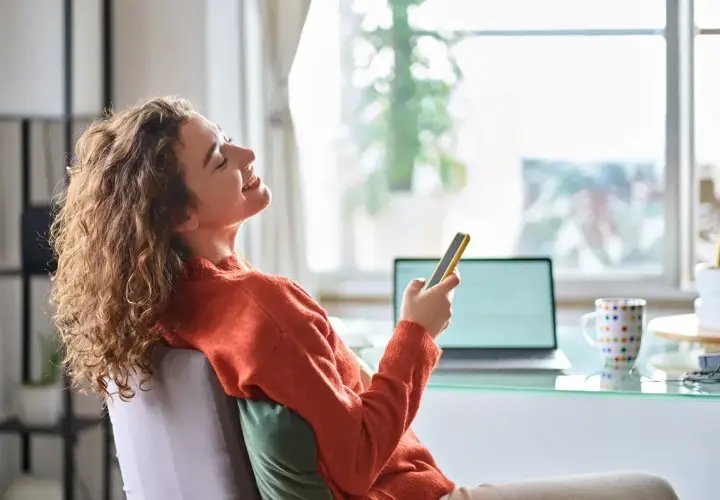 Kobieta z kręconymi włosami śmieje się, trzymając telefon, siedząc przy biurku z laptopem.