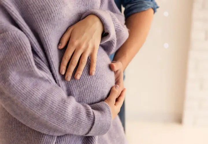 Dłoń kobiety w fioletowym swetrze obejmuje ciążowy brzuch, obok dłoń partnera.