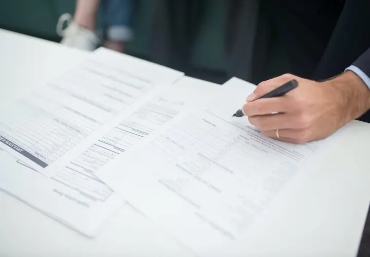 Dłoń z długopisem podpisująca dokumenty na białym stole.