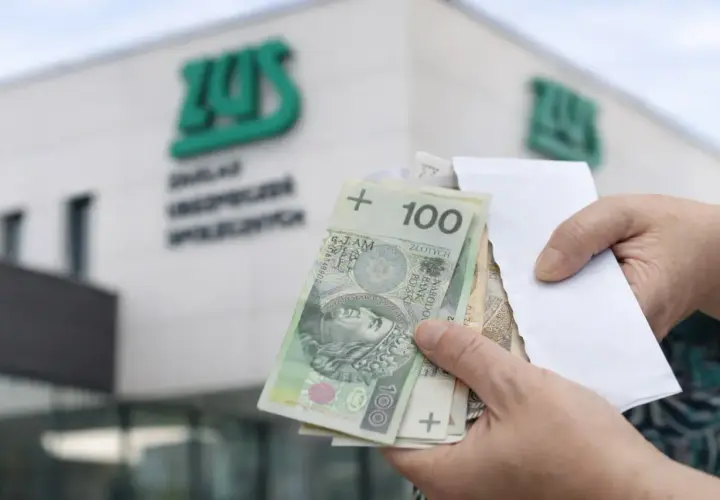 Dłonie trzymające polskie banknoty 100 zł przed budynkiem ZUS.