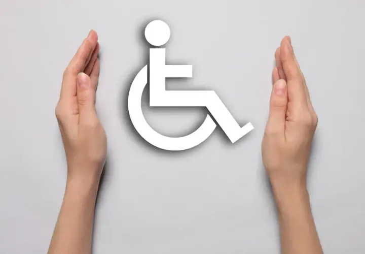 Dwie dłonie otaczają biały symbol osoby na wózku inwalidzkim na jasnoszarym tle.