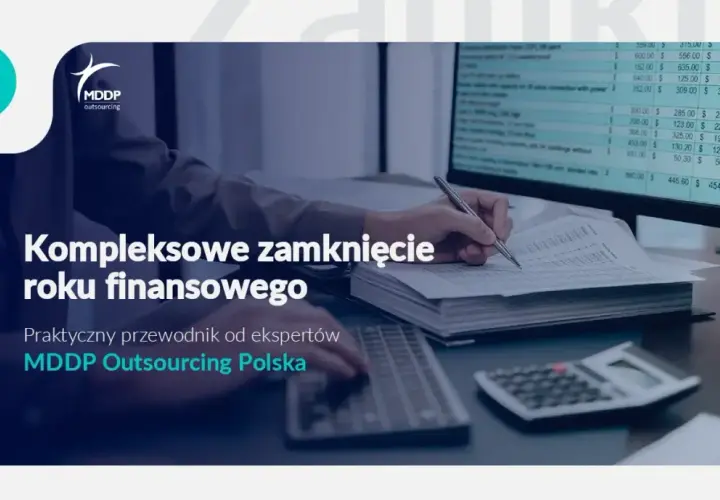 Osoba pisząca ręcznie na dokumentach przy biurku z komputerem i kalkulatorem.