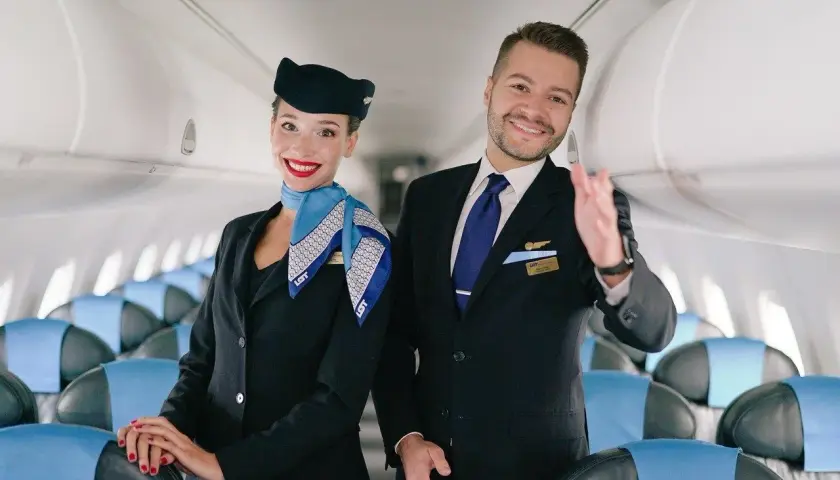 Uśmiechnięta stewardessa i steward w mundurach LOT stoją w samolocie, na tle rzędów siedzeń.