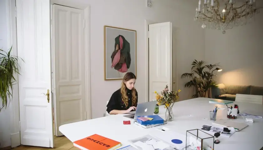 Kobieta pracuje przy laptopie w jasnym pomieszczeniu z obrazem na ścianie i bukietem kwiatów na stole.