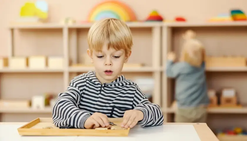Chłopiec w bluzie w paski skupiony na zadaniu z materiałami Montessori. W tle inne dziecko przy półce.