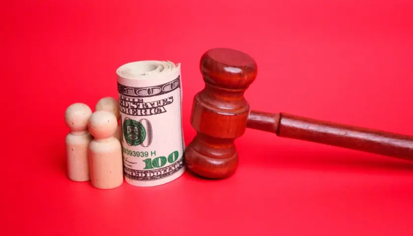 Drewniany młotek sędziowski, zwinięty banknot 100 dolarów i trzy drewniane figurki symbolizujące rodzinę. Pytanie: do kiedy alimenty na dziecko?