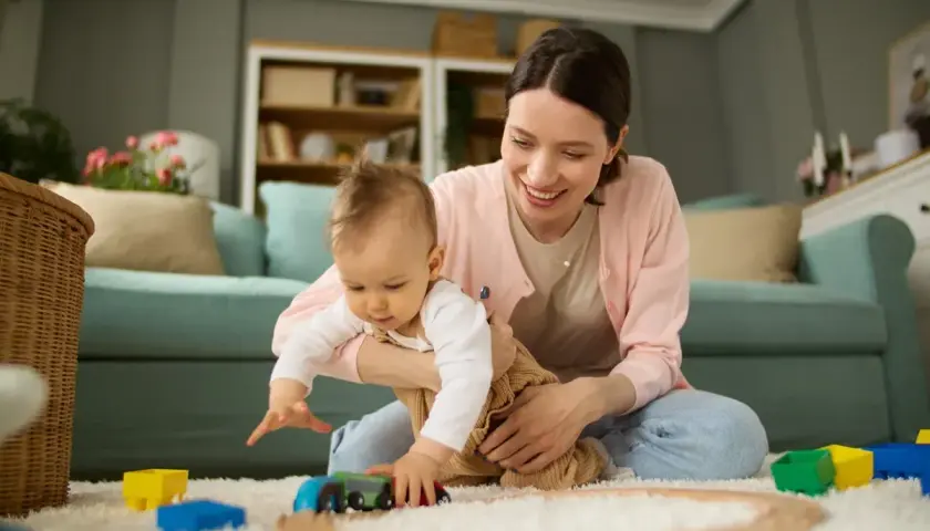 Uśmiechnięta mama bawi się z dzieckiem klockami i pociągiem na dywanie w salonie.
