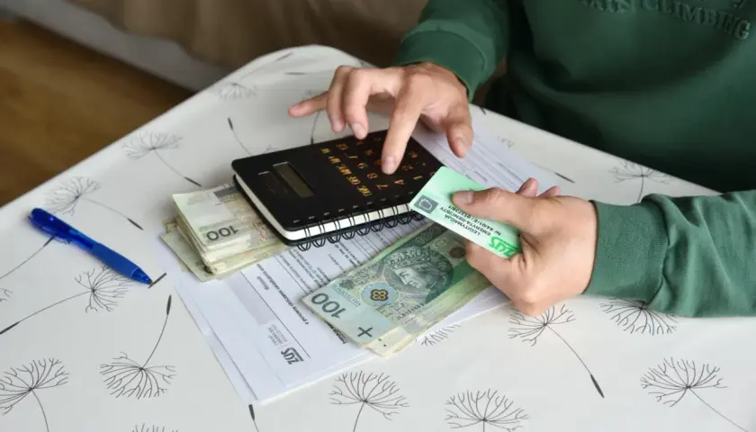 Dłonie trzymające pieniądze, kalkulator i dokumenty ZUS na stoliku.