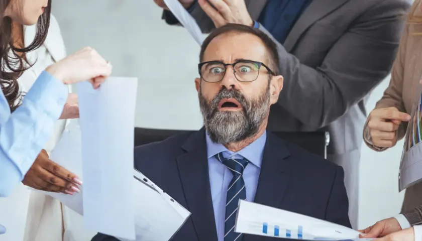 Przerażony mężczyzna w garniturze otoczony przez ludzi z dokumentami.