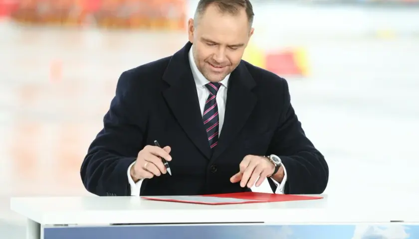 Mężczyzna w garniturze i płaszczu podpisuje dokumenty przy stole.