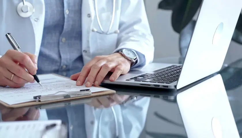 Lekarz w białym kitlu pisze na dokumentach, obok laptop.