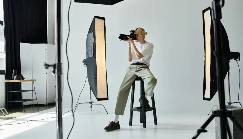 Fotograf siedzi na stołku w studiu, trzymając aparat i robiąc zdjęcie.