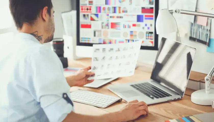 Mężczyzna przy biurku przegląda wydruki, przed nim komputer z kolorowymi grafikami i laptop.