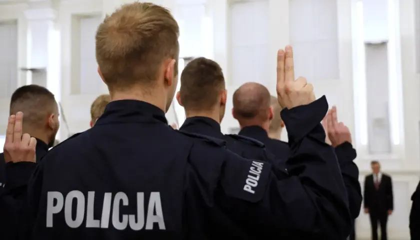 Policjanci w mundurach z napisem "POLICJA" podnoszą ręce z dwoma palcami uniesionymi do góry.