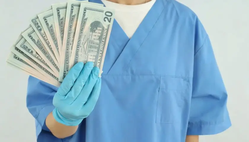 Higienistka stomatologiczna w niebieskim fartuchu trzyma wachlarz banknotów 20-dolarowych.