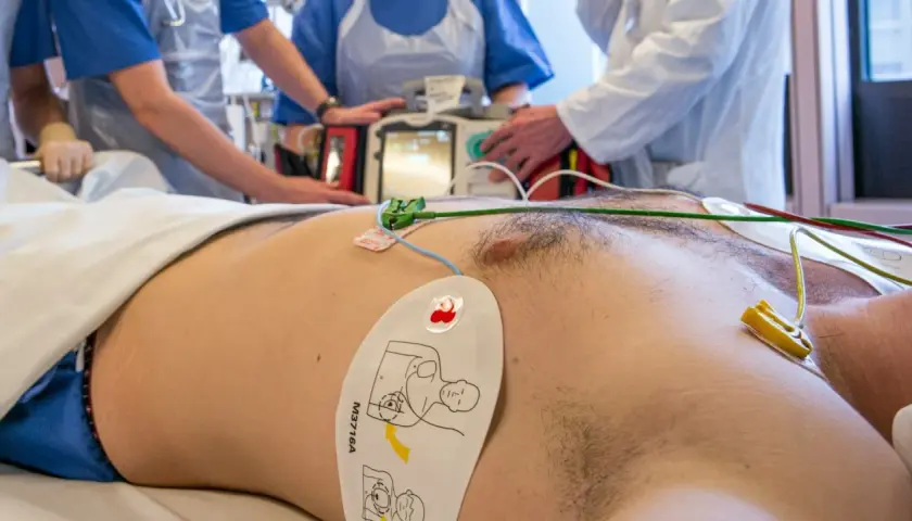 Pacjent z elektrodami EKG i defibrylatorem w tle, otoczony przez personel medyczny.