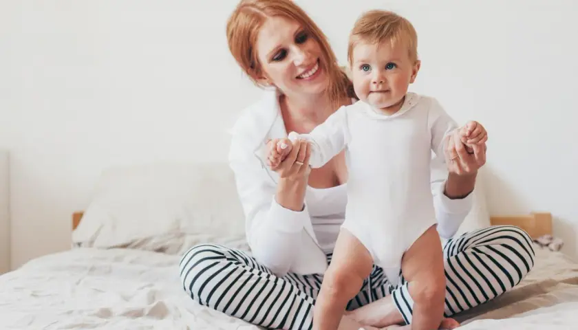 Uśmiechnięta mama z rudymi włosami pomaga 8-miesięcznemu dziecku w białym body wstać na łóżku.