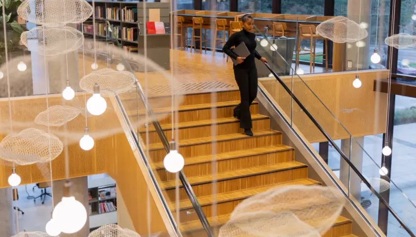 Kobieta z laptopem schodzi po drewnianych schodach w nowoczesnej bibliotece z ozdobnymi chmurkami i lampami.