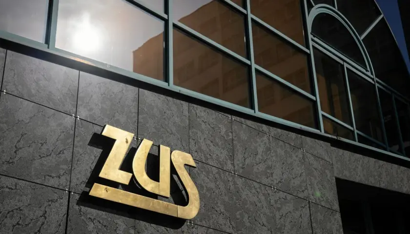 Złote logo ZUS na ciemnej, kamiennej elewacji budynku, nad nim okna z odbiciem.