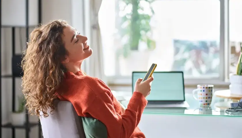 Kobieta z kręconymi włosami śmieje się, trzymając telefon, siedząc przy biurku z laptopem.
