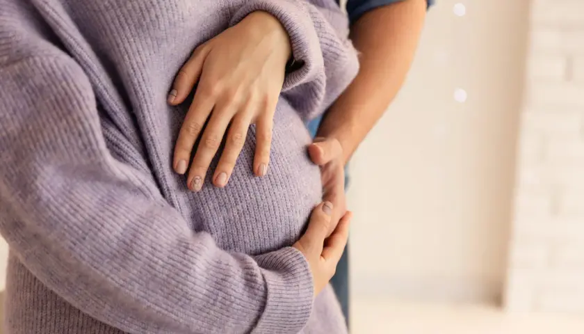 Dłoń kobiety w fioletowym swetrze obejmuje ciążowy brzuch, obok dłoń partnera.