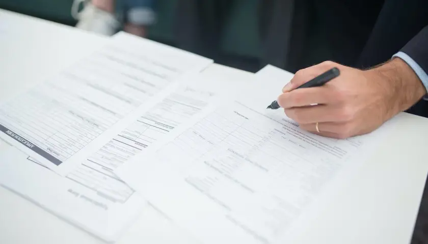 Dłoń z długopisem podpisująca dokumenty na białym stole.