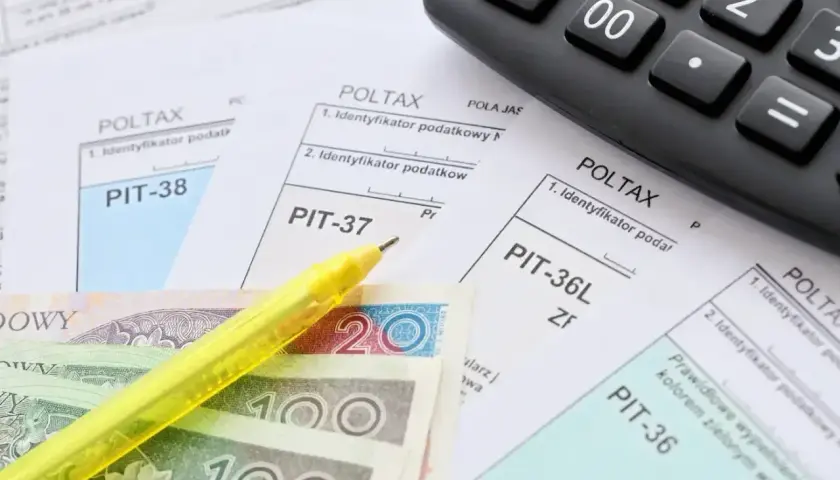 Formularze PIT, polskie banknoty, żółty długopis i kalkulator.