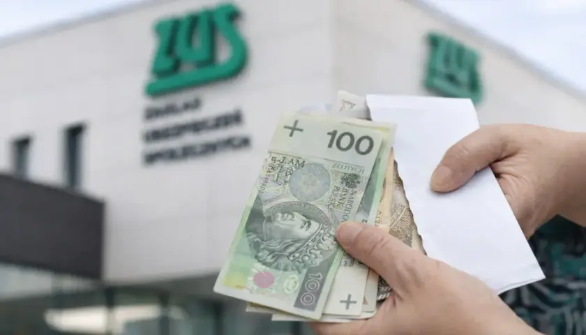 Dłonie trzymające polskie banknoty 100 zł przed budynkiem ZUS.