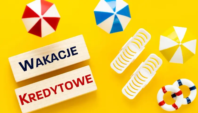Drewniane klocki z napisem "Wakacje Kredytowe" na żółtym tle, otoczone miniaturowymi parasolkami plażowymi i kołami ratunkowymi.