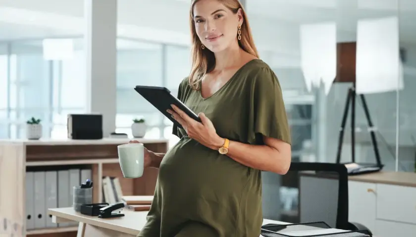 Ciężarna kobieta w zielonej sukience trzyma tablet i kubek w biurze.